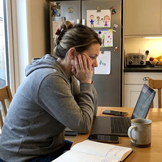 A woman at a kitchen table working on a laptop, her hand holding the side of her face